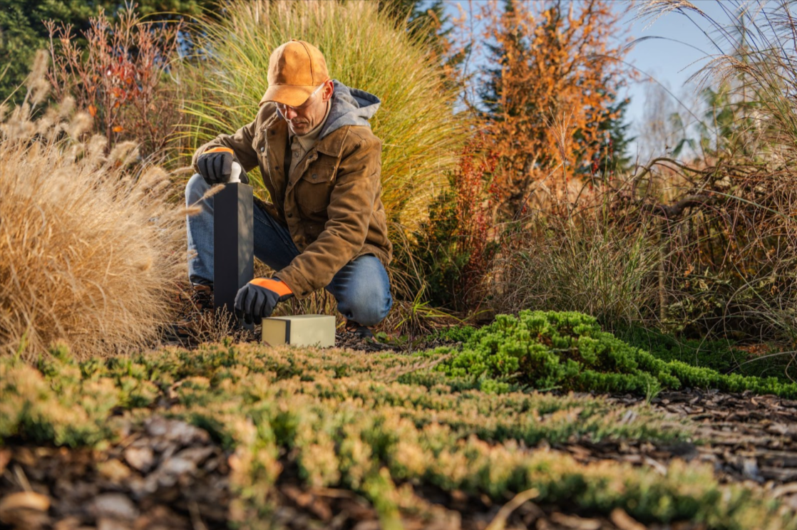 An image demonstrating what landscapers do. A landscaper wearing an orange baseball cap, brown jacket, hoodie, jeans, and gloves kneeling, tending to low-maintenance plants.