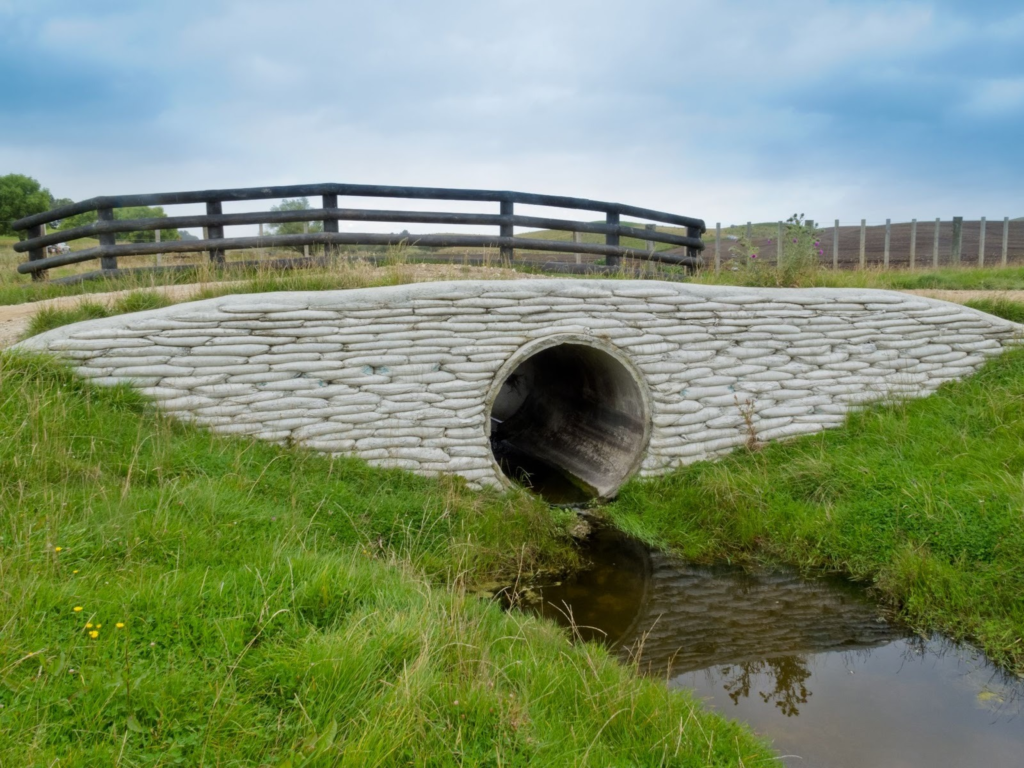 A storm culvert under a white stone bridge on a green field.