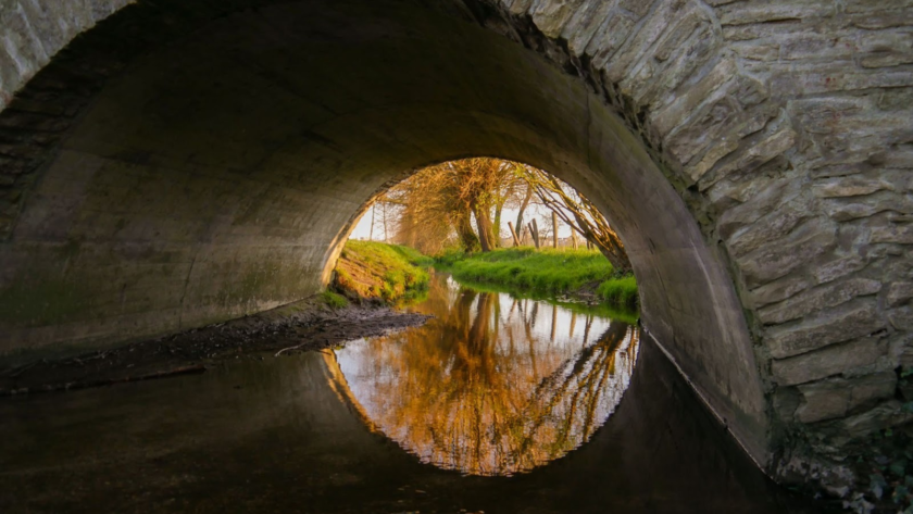 A large stone arch culvert under a stone bridge above a calm river.