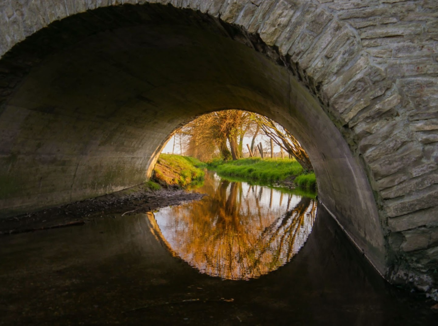 A large stone arch culvert under a stone bridge above a calm river.