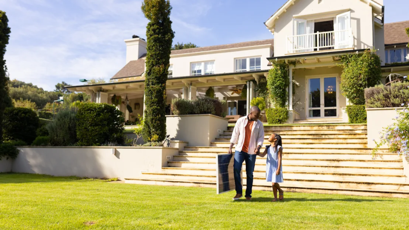 A father and daughter hold hands and walk over their front lawn.